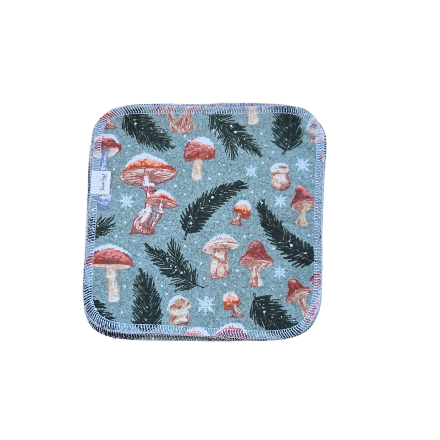 A square cloth napkin with a Christmas-themed pattern featuring mushrooms and pines on a green background, placed on a wooden surface.