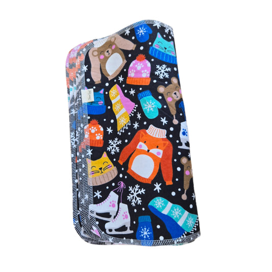 A multicolored towel with a winter-themed design, featuring bears, snowflakes, and other cold-weather clothing items, laid out on a wooden surface.
