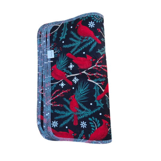 A square-shaped, black cloth towel with a Christmas-themed design featuring red cardinals and green foliage, placed on a wooden surface.