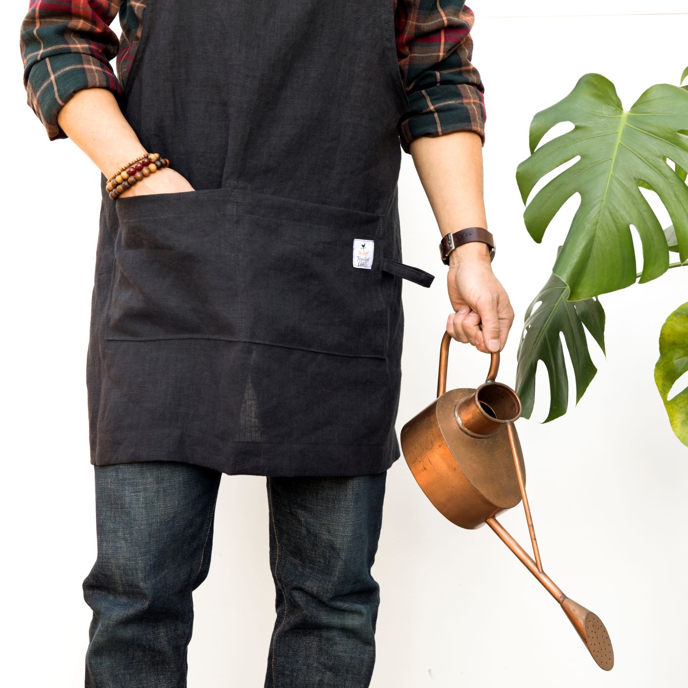 Photo of a man holding a vintage watering can, next to a monstera plant wearing vintage bead bracelets and modeling a black linen apron by farm girl cloth co.