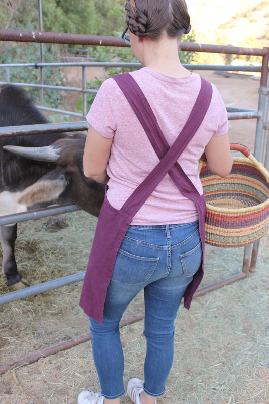 Photo of a woman standing next to a cow, holding a woven basket and showing the crossback design of the farm girl cloth co linen apron